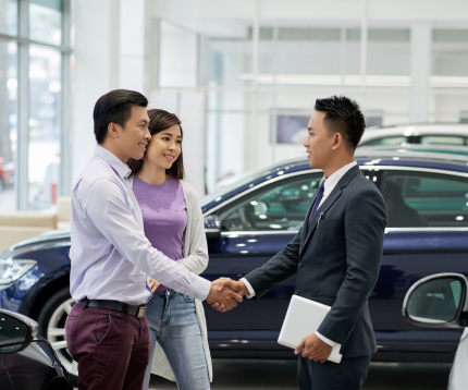 Smiling couple shaking hands with a car salesman inside a car dealership.
